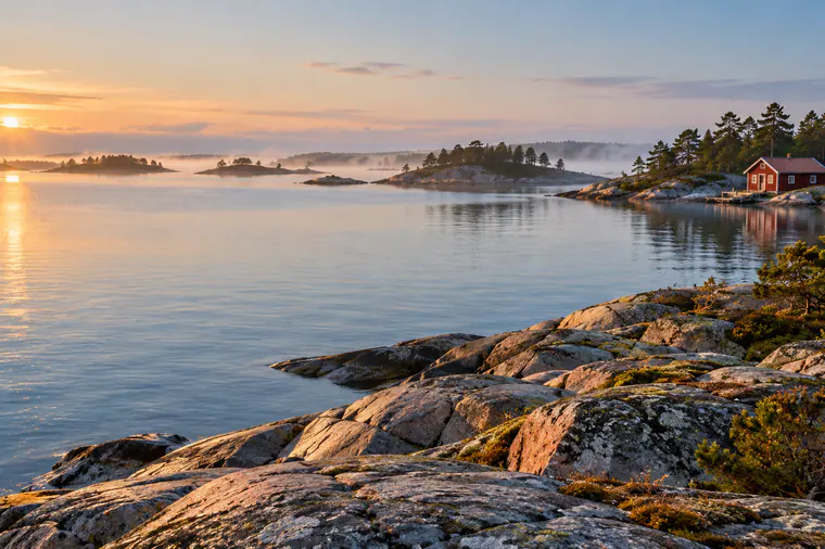 Klippor och tallar i skärgården i morgonljus, med en röd stuga vid vattnet.